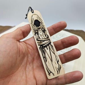 Könnte beinhalten: Ein Lesezeichen aus Holz mit einem Design einer Kapuzenfigur und einer schwarzen Kordel. Das Bild ist in das hellfarbene Holz eingebrannt und erzeugt einen Kontrast zwischen dem dunklen Design und der natürlichen Holzmaserung. Das Lesezeichen wird in einer Hand gehalten.