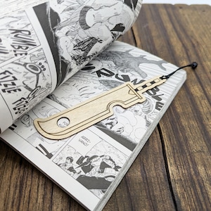 May include: A wooden bookmark shaped like a sword, resting on the pages of a manga book. The bookmark is light brown with etched details and a black string. The manga pages are black and white.