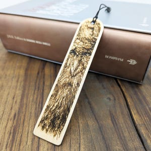 May include: A wooden bookmark with a detailed pyrography design of an owl's face. The bookmark is rectangular with a rounded top and a black string. The owl's features are intricately burned into the light-colored wood, creating a striking contrast.