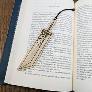 Peut inclure: Marque-page en bois en forme d'épée, avec des détails gravés. Le marque-page est brun clair et possède un cordon noir. Il est posé sur les pages d'un livre ouvert.
