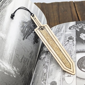 May include: A wooden bookmark shaped like a sword, with a black cord attached. The bookmark is light brown with a textured gold center and is placed in an open book with black and white illustrations.