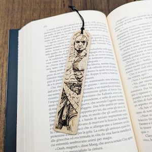Può includere: Un segnalibro in legno con un'incisione di un guerriero calvo con un'espressione seria. Il segnalibro ha un cordino nero ed è posto all'interno di un libro aperto.