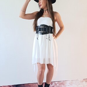 Puede incluir: Una mujer con sombrero negro, vestido blanco y cors&eacute; negro. El vestido tiene un dobladillo asim&eacute;trico. Tambi&eacute;n lleva botas de plataforma negras. El cors&eacute; tiene hebillas y lazos decorativos.
