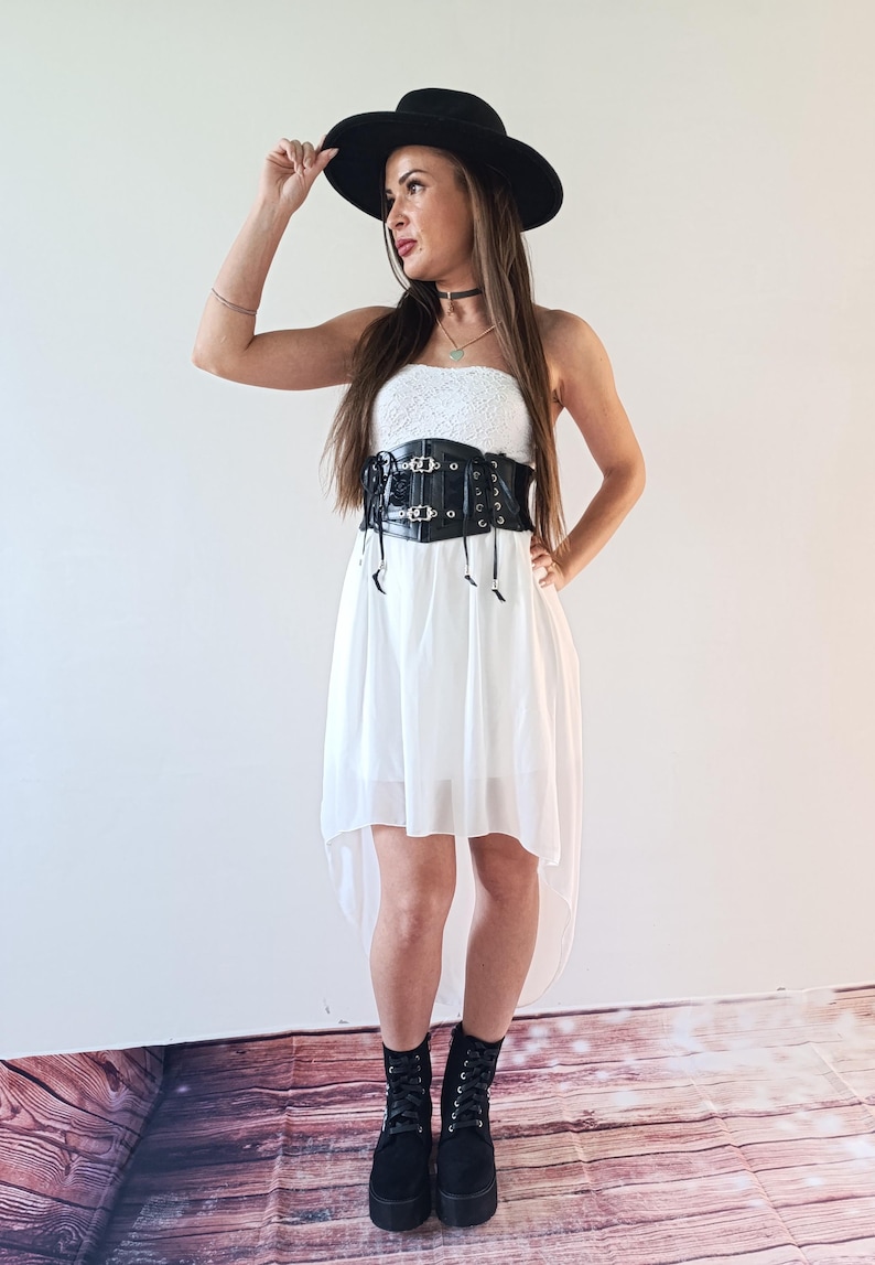 Puede incluir: Una mujer con sombrero negro, vestido blanco y cors&eacute; negro. El vestido tiene un dobladillo asim&eacute;trico. Tambi&eacute;n lleva botas de plataforma negras. El cors&eacute; tiene hebillas y lazos decorativos.