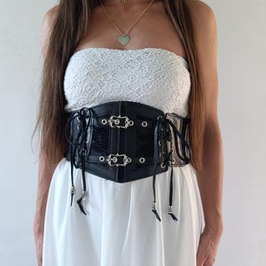 Puede incluir: Un cors&eacute; negro con hebillas y ojales plateados, usado sobre un top de encaje blanco y un vestido blanco. El cors&eacute; tiene un acabado brillante y se ata con cordones en la parte delantera. Tambi&eacute;n se ven un collar negro y un collar dorado con un colgante de coraz&oacute;n.
