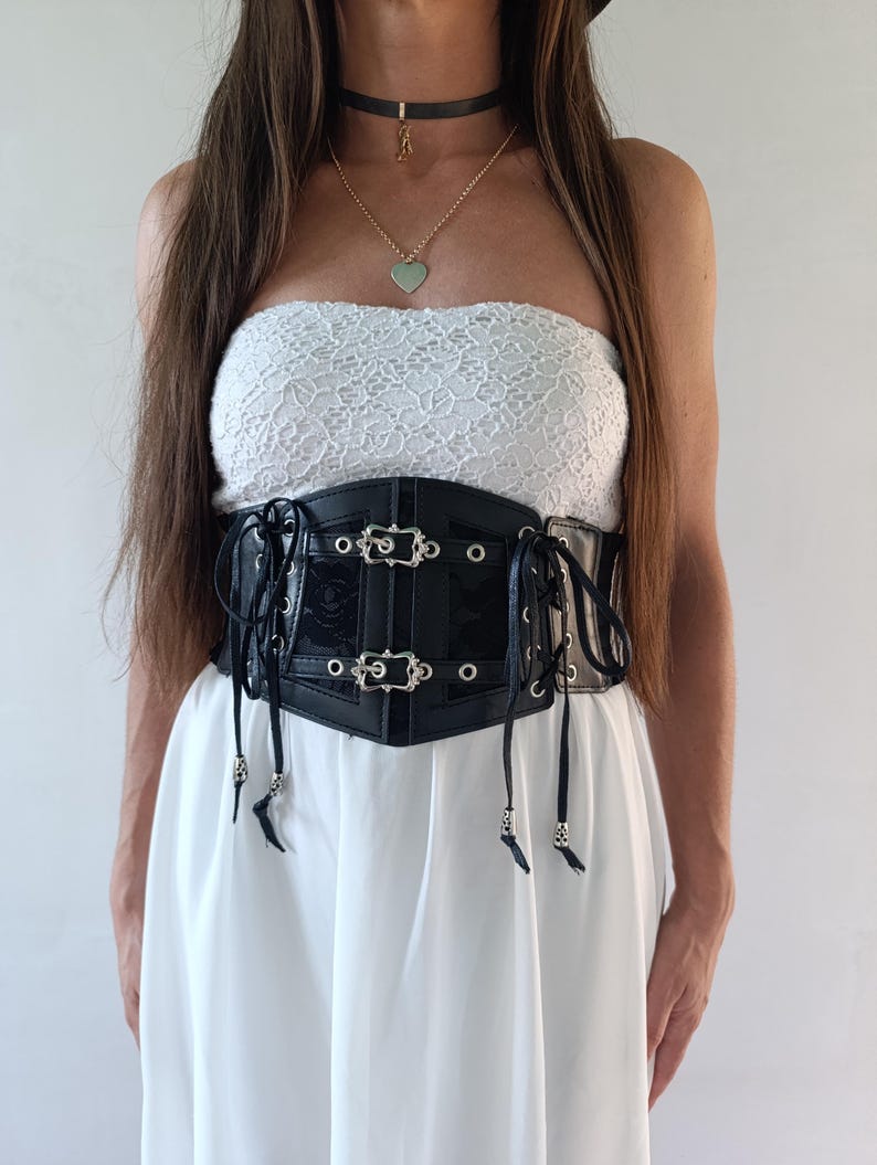 Puede incluir: Un cors&eacute; negro con hebillas y ojales plateados, usado sobre un top de encaje blanco y un vestido blanco. El cors&eacute; tiene un acabado brillante y se ata con cordones en la parte delantera. Tambi&eacute;n se ven un collar negro y un collar dorado con un colgante de coraz&oacute;n.