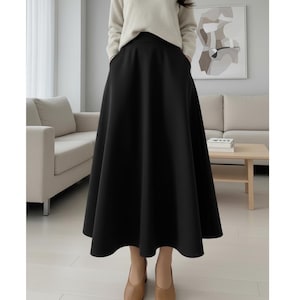 Puede incluir: Una falda negra acampanada de cintura alta. La falda se combina con un suéter color crema y zapatos color canela. La modelo está de pie en una habitación bien iluminada con muebles de tonos neutros.