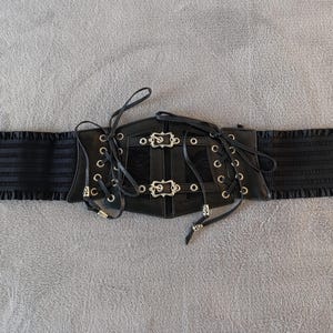Puede incluir: Cintur&oacute;n cors&eacute; negro con hebillas y ojales plateados. El cintur&oacute;n presenta un dise&ntilde;o con cordones en la parte delantera y paneles laterales el&aacute;sticos. Un accesorio de moda con elementos decorativos para varios atuendos.
