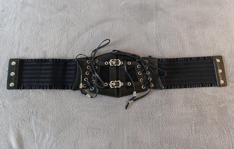 Puede incluir: Cintur&oacute;n cors&eacute; negro con hebillas y ojales plateados. El cintur&oacute;n presenta un dise&ntilde;o con cordones en la parte delantera y paneles laterales el&aacute;sticos. Un accesorio de moda con elementos decorativos para varios atuendos.