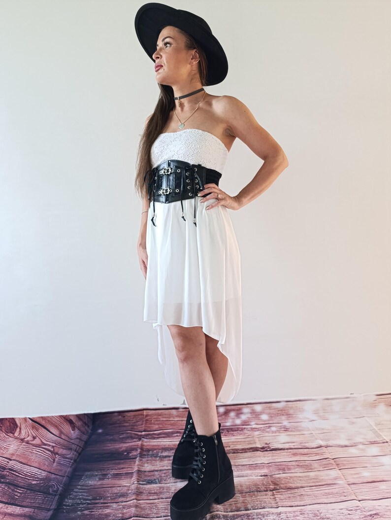 Puede incluir: Un atuendo femenino con un sombrero negro de ala ancha, un vestido blanco sin tirantes con corpi&ntilde;o de encaje y un cintur&oacute;n cors&eacute; negro. El conjunto se completa con botas de plataforma negras. El vestido tiene un dobladillo alto-bajo.