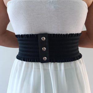 Puede incluir: Un cintur&oacute;n el&aacute;stico negro con un ribete de volantes decorativo y tres botones plateados. El cintur&oacute;n se usa sobre un top blanco sin tirantes y un vestido blanco, creando una silueta de cintura ce&ntilde;ida.