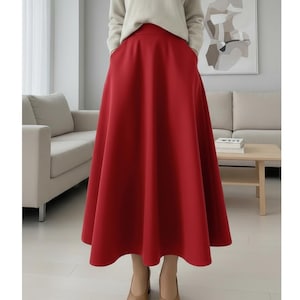 May include: A full-length shot of a red skirt with a wide A-line silhouette. The skirt has pockets and falls to the mid-calf. The person is wearing a cream-colored sweater and tan shoes. The background is a neutral-toned room.