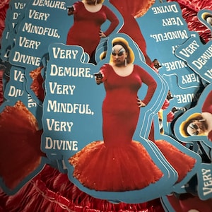 Puede incluir: Un montón de pegatinas azules con una drag queen con un vestido rojo y una pistola. Las pegatinas tienen las palabras "VERY DEMURE, VERY MINDFUL, VERY DIVINE". Las pegatinas están sobre una superficie roja texturizada.
