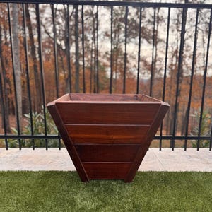 May include: A square, wooden planter with a rich, dark brown finish. The planter has a tapered design, with horizontal wooden slats forming the sides. It sits on a green, grassy surface, with a black metal railing and trees in the background.
