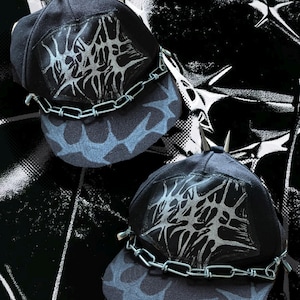 May include: Two black baseball caps featuring silver metal spikes on the crown and a silver chain across the brim. Each cap displays a large, intricate silver graphic patch on the front panel and a dark blue, abstract, web-like pattern on the brim.