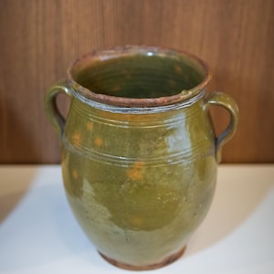 Puede incluir: Una olla de cerámica verde con dos asas. La olla tiene una abertura ancha y una superficie ligeramente texturizada. Presenta líneas horizontales y motas anaranjadas. El borde es de color marrón oscuro. La olla probablemente se utiliza para almacenamiento o exhibición.