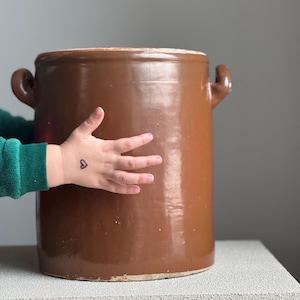 Peut inclure: Un grand pot en céramique marron cylindrique avec deux petites poignées. Le pot a une finition brillante et un bord de couleur claire. La main d'un enfant avec un tatouage de cœur touche le pot.