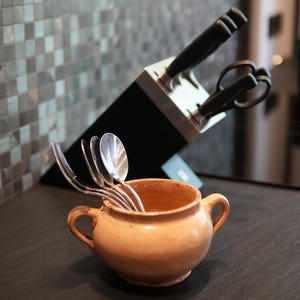 Könnte beinhalten: Ein kleiner, hellbrauner Keramiktopf mit zwei Henkeln, gefüllt mit silbernen Löffeln. Dahinter befindet sich ein schwarzer Messerblock mit Küchenmessern und einer Schere. Der Hintergrund ist eine Mosaikfliesenwand.