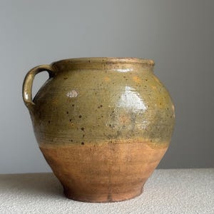 May include: An antique ceramic pot with a single handle. The upper section is a mottled olive green, fading into a warm, earthy brown at the base. The pot has a rounded body and a slightly flared rim, displaying a vintage aesthetic. It is approximately 25 cm in height.