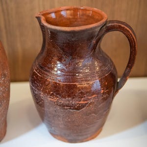 Może przedstawiać: Brązowy ceramiczny dzban z uchwytem i wylewką. Dzban ma teksturowaną powierzchnię i ciemnobrązowy kolor z jaśniejszymi brązowymi akcentami. Dzban jest prawdopodobnie ręcznie robiony i ma rustykalny wygląd.