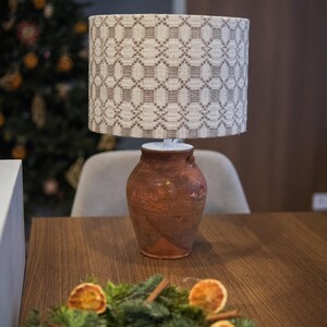 May include: A decorative table lamp with a patterned lampshade and a rustic, terracotta-colored base. The lamp sits on a wooden table, with a festive wreath of greenery, dried oranges, and cinnamon sticks in the foreground. A Christmas tree is visible in the background.