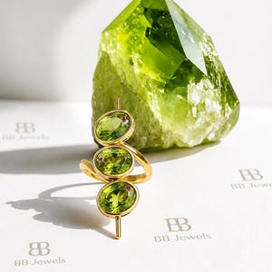 Op de afbeelding: Een gouden ring met drie verticaal uitgelijnde ovaal geslepen groene peridoot edelstenen, elk geaccentueerd met een klein roze steentje. De ring staat voor een grote, ruwe, doorschijnende limoengroene kristalpunt. De tekst BB Jewels is zichtbaar.