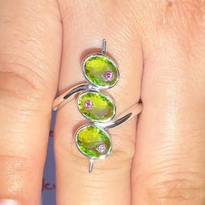 May include: A silver ring featuring three vertically aligned oval-shaped lime green stones, each accented with a small pink dot in the center. The band is thin and polished, wrapping around the finger to support the trio of green settings.