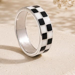 May include: A silver-toned band ring featuring a repeating checkerboard pattern of alternating black and white square enamel inlays around the entire outer circumference of the metal band, creating a high-contrast geometric design.