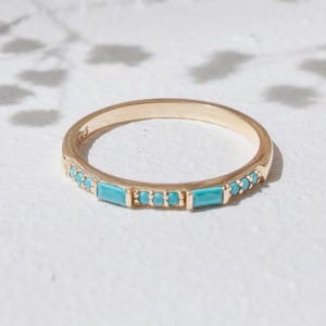 Op de afbeelding: Een goudkleurige ring met een band van rechthoekige turkooizen stenen. De ring staat tegen een witte achtergrond met zachte schaduwen en kleine bloemen.