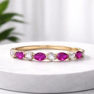 Può includere: Un anello a fascia in oro con pietre preziose rosa vivaci a taglio ovale e pietre preziose chiare rotonde alternate. L'anello è esposto su una superficie bianca, mostrando il suo design elegante e la sua brillantezza.