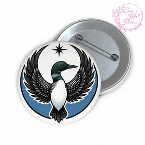 May include: A round button with a graphic of a loon with outstretched wings, a star, and a blue border. The loon has black and white feathers, a green head, and a black beak. The button is white with a metal pin back. The text "Lina Rebel Bloom" is in the upper right corner.