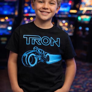 Könnte beinhalten: Schwarzes T-Shirt mit dem Wort "TRON" in blauer Neon-Schrift und einer blauen Lightcycle-Grafik. Das T-Shirt wird von einem lächelnden Kind in blauen Jeans getragen.