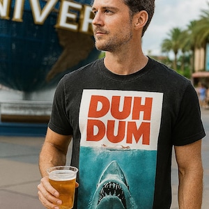 May include: Black t-shirt with a graphic featuring the words "DUH DUM" in red letters above an image of a shark in the ocean. The person is holding a clear plastic cup with a light-colored beverage.