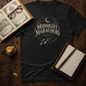 Può includere: T-shirt nera con un design circolare. Il design presenta le parole "Midnight Marauders" sopra impronte, una luna crescente e alberi. Il testo recita "After Dark Only - Est. 1971". La maglietta è esposta con libri e una candela.