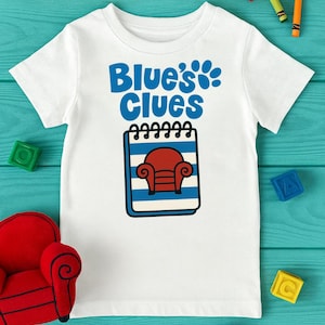 May include: White t-shirt featuring the text "Blue's Clues" in blue, along with a paw print and a notepad with a red chair. The shirt is surrounded by colorful blocks and crayons, creating a playful and engaging design.