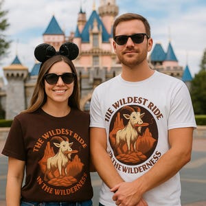 Op de afbeelding: Een vrouw draagt een bruin t-shirt en een man een wit t-shirt, beide met de tekst "THE WILDEST RIDE IN THE WILDERNESS". De afbeelding toont een geit met een frisbee, tegen een woestijnlandschap. Ze dragen zonnebrillen.
