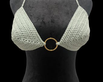 May include: A sage green crochet halter top with a triangle cup design. The top is connected by a gold-colored ring in the center. The halter straps are thin and made of the same crochet material. The top is displayed on a black mannequin.
