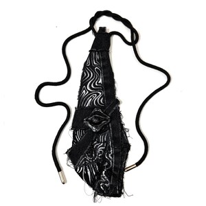 Peut inclure: Un collier textile noir au design abstrait unique. Le pendentif présente un motif texturé avec des accents blancs et est relié à un cordon noir. Le collier est doté d'un fermoir en métal argenté.