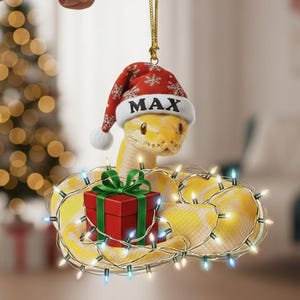 Könnte beinhalten: Ein gelber Schlangen-Weihnachtsschmuck mit einem Weihnachtsmannhut mit dem Namen "MAX". Die Schlange ist um eine rote Geschenkbox mit grüner Schleife gewickelt und von Lichterketten umgeben. Eine festliche Weihnachtsdekoration.