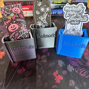 May include: Three 3D-printed plastic holders with the word Bookmarks embossed on the front. The holders are purple, black, and blue, each containing a decorative bookmark. The bookmarks feature Halloween patterns, a floral coffin design, and text.