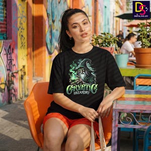 May include: Black t-shirt featuring a wizard graphic with the text "Cryptic Wizardry" in a stylized font. The wizard is depicted with a long beard and a pointed hat, surrounded by green smoke. The shirt is worn by a person sitting outdoors.