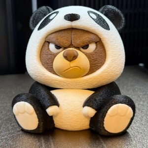 May include: A figurine of a bear wearing a panda costume. The bear has a grumpy expression, with a brown face and a white and black panda hood. The bear is sitting with its paws out, and the figurine is on a gray surface.