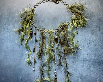 May include: A necklace featuring a circular bronze-toned chain adorned with hanging strands of olive green fringe. The design includes small metallic leaf charms and multi-colored beads attached to thin chains, creating a layered, textured appearance.