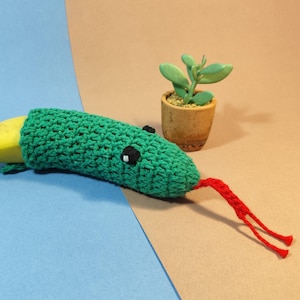 Op de afbeelding: Een banaan met een groen gehaakt slangenkostuum met zwarte ogen en een rode tong. Op de achtergrond staat een kleine vetplant in een pot. De banaan ligt op een blauw en bruin oppervlak.