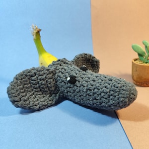 Op de afbeelding: Een handgemaakt grijs gehaakt olifantenspeelgoed met zwart-witte ogen, een gele banaan en een kleine vetplant in pot. De olifant is het middelpunt, tegen een blauw en bruin achtergrond.