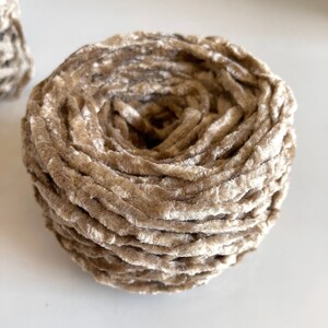 May include: A skein of beige velvet yarn, wound into a circular shape. The yarn is soft and fluffy, with a subtle sheen.