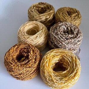 May include: A collection of six yarn skeins in various shades of gold, brown, and beige. The yarn textures vary, with some appearing smooth and others having a looped, textured appearance. The skeins are arranged on a white surface.