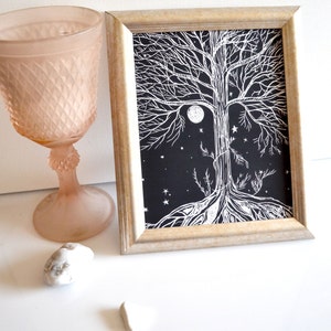 Puede incluir: Una impresi&oacute;n enmarcada en blanco y negro de un &aacute;rbol con ra&iacute;ces y ramas que se extienden hasta los bordes del papel. El &aacute;rbol est&aacute; rodeado de una luna llena y estrellas. La impresi&oacute;n est&aacute; en un marco de madera claro.