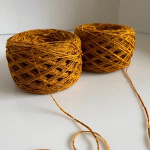 May include: Two spools of gold colored yarn, one is partially unwound with a long strand of yarn extending from it.
