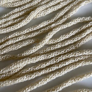May include: Close-up of several strands of off-white, crocheted rope. The rope appears to be made of a natural fiber, with a slightly textured surface. The strands are arranged diagonally across the frame, creating a sense of depth and texture. The overall aesthetic is simple and rustic.